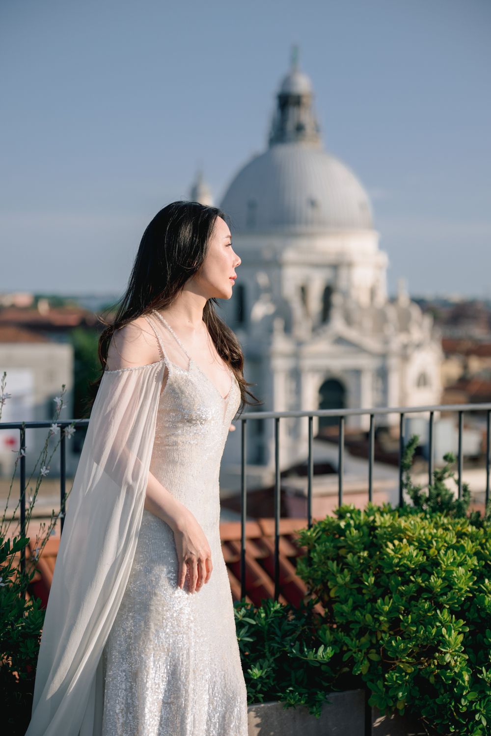 Bride in white sequin gown with sheer cape stands on rooftop terrace with Santa Maria della Salute dome, Venice.
