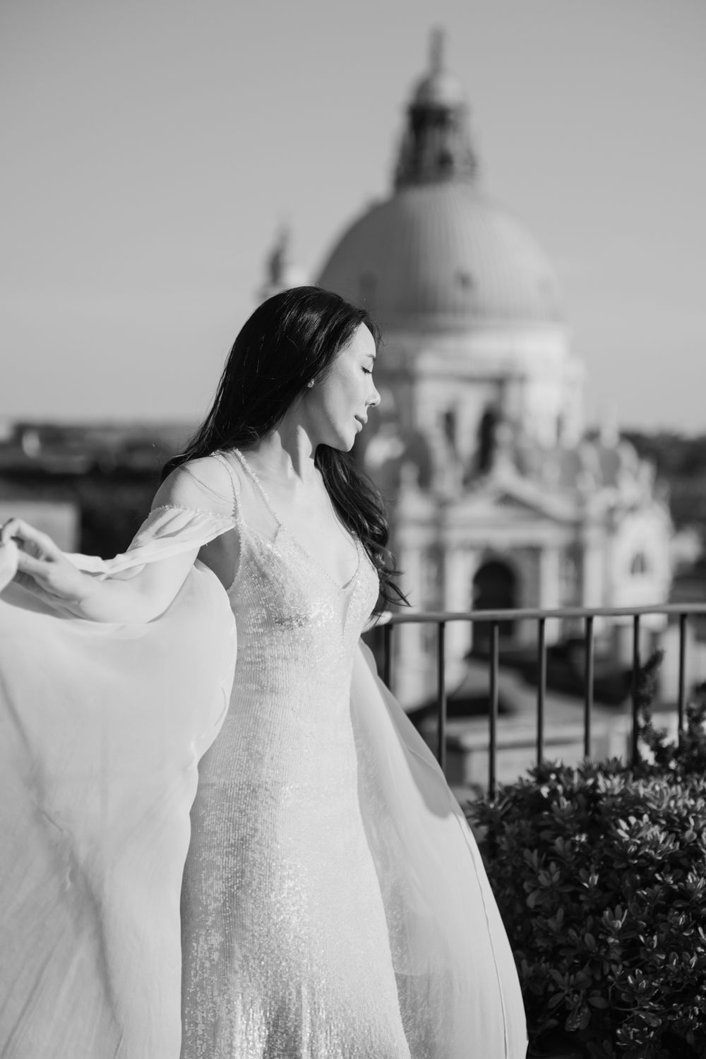 Black and white photo of a woman in a flowing white dress on a balcony with Santa Maria della Salute in Venice behind her.
