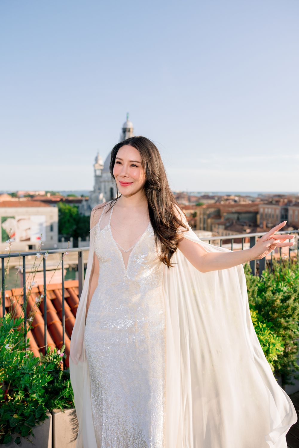 A bride in a sequined white gown with flowing cape poses on a rooftop terrace overlooking a European cityscape.