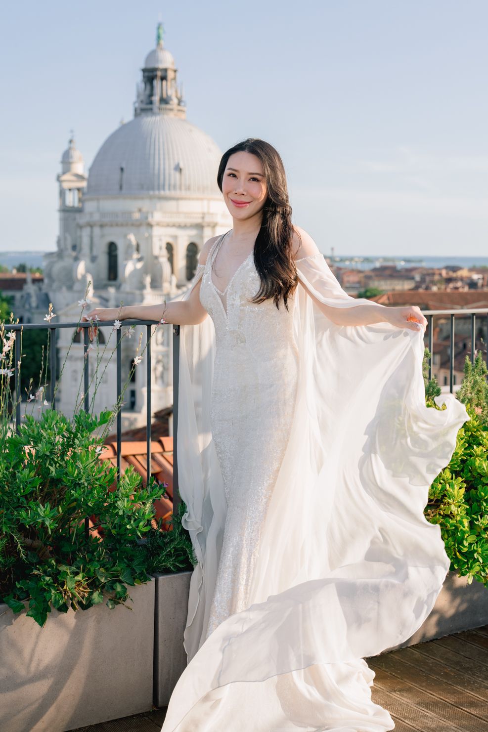 Bride in flowing white gown poses on rooftop terrace with Venice's Santa Maria della Salute dome backdrop.