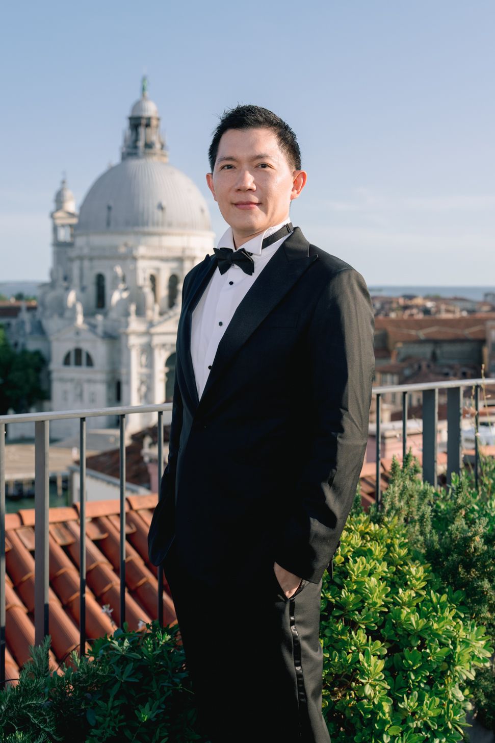 A man in a black tuxedo poses on a rooftop terrace with the Santa Maria della Salute basilica in Venice behind him.