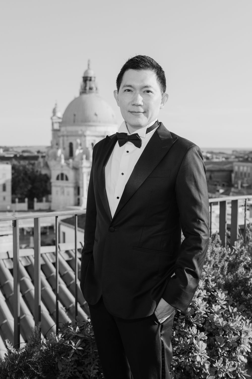 A man in a tuxedo poses on a rooftop terrace with a domed cathedral and cityscape in the background.