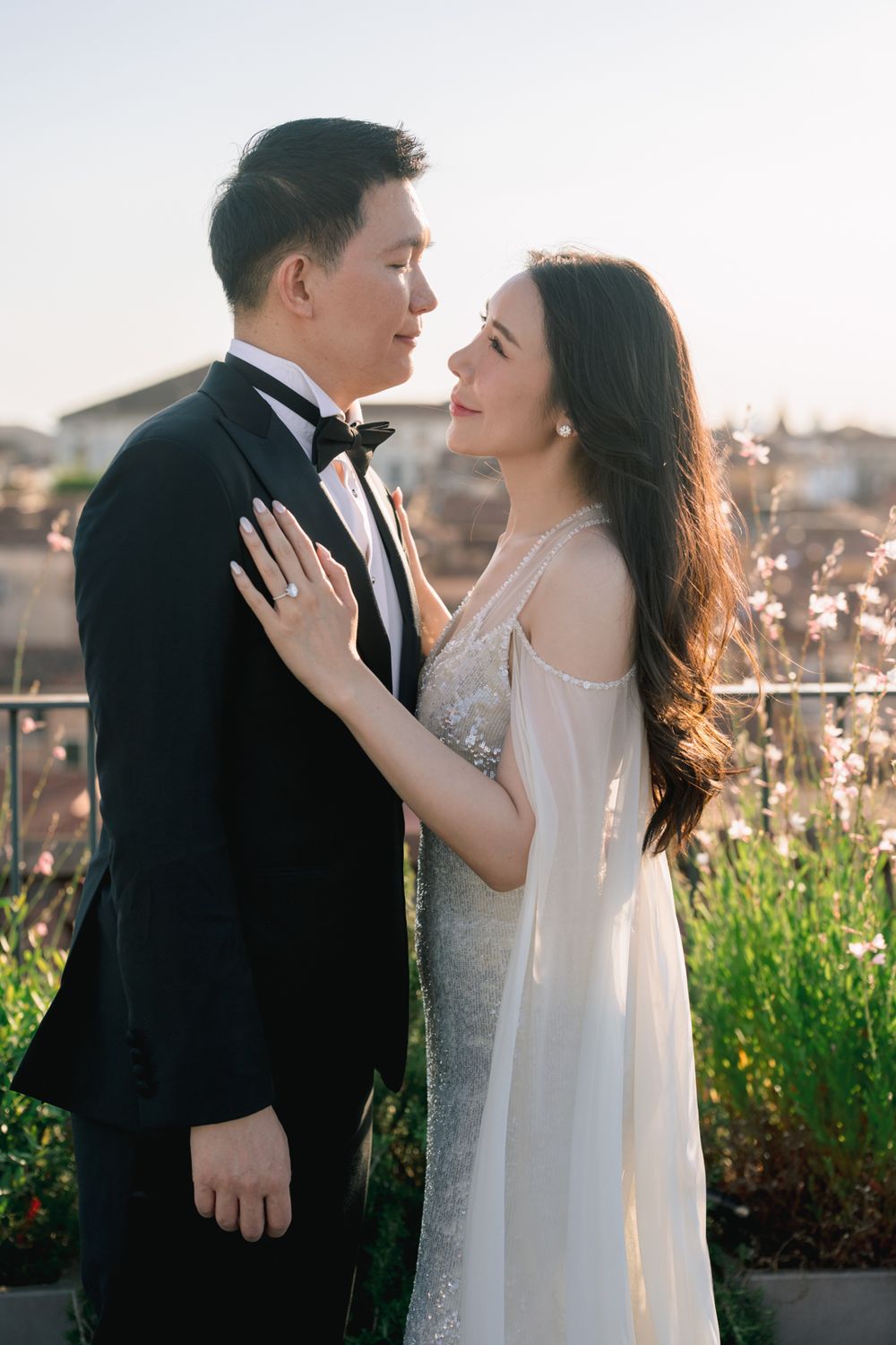 A couple in elegant wedding attire share a romantic moment outdoors, the bride in white gown and groom in black tuxedo.