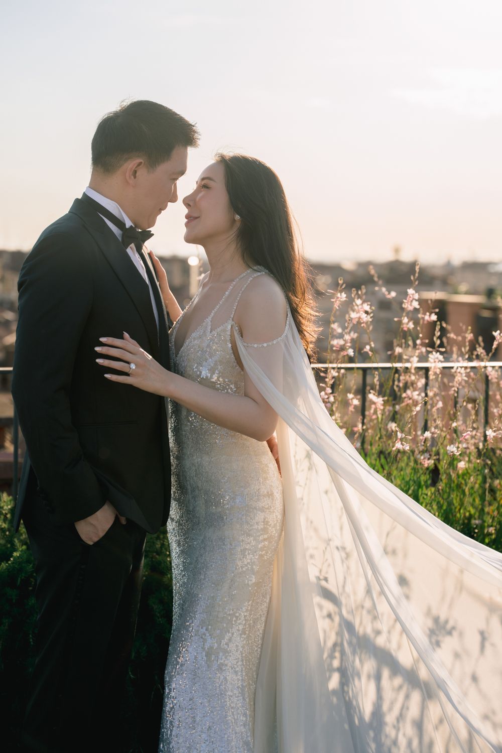 Couple in wedding attire embracing on a rooftop at sunset, bride in sequined gown with flowing veil, groom in black tuxedo.