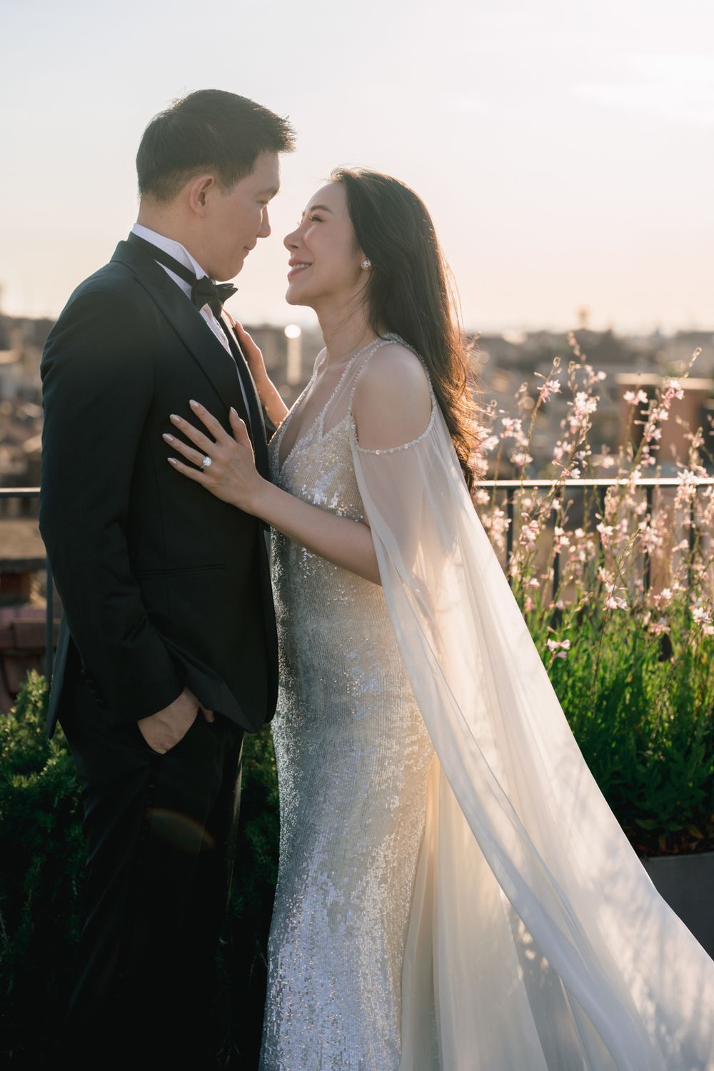 Bride in lace gown and veil smiles at groom in black tuxedo during golden hour rooftop wedding portrait.
