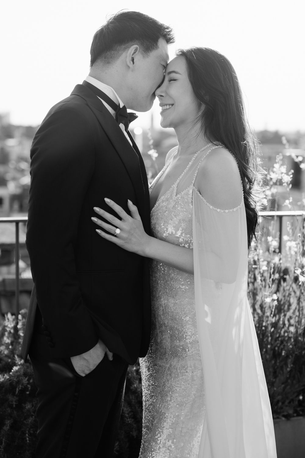 Black and white photo of a smiling bride in a sequin gown and groom in a tuxedo sharing a romantic moment outdoors.