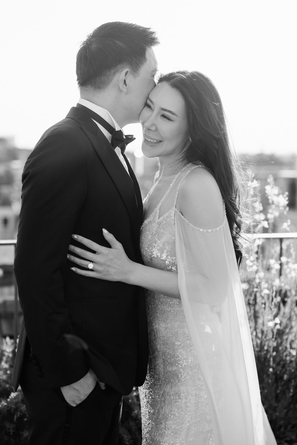 Black and white photo of a smiling bride in sequin gown with groom in tuxedo sharing a tender moment outdoors.