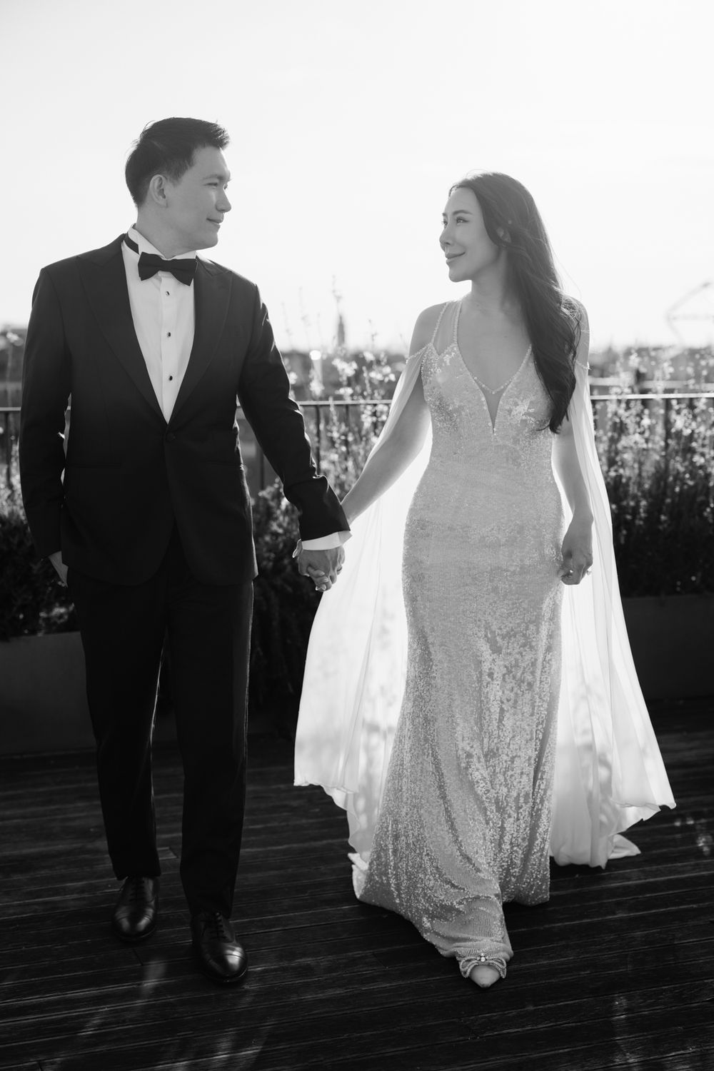Black and white photo of a couple in wedding attire holding hands and walking on a rooftop deck.