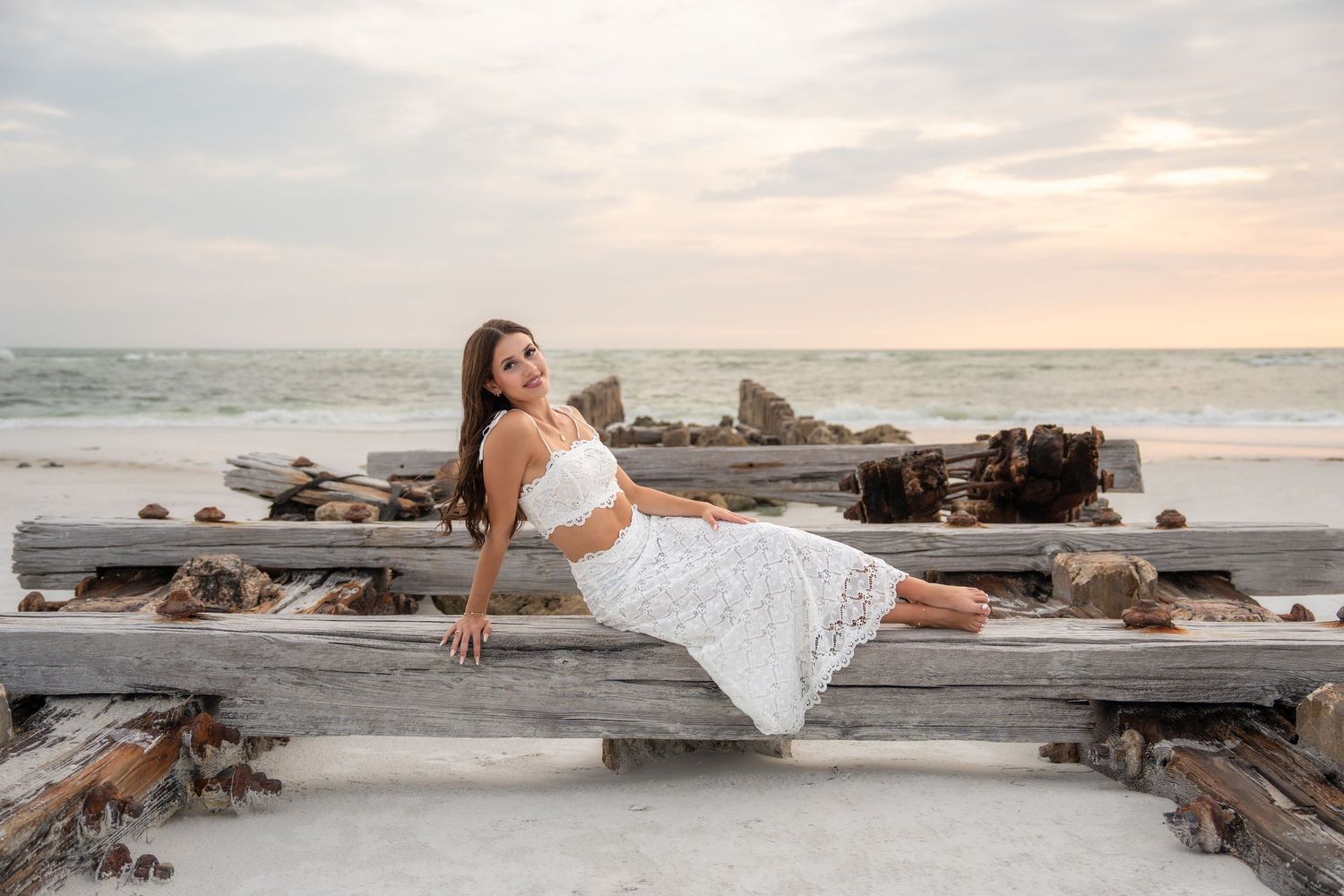 Woman in white crop top and skirt relaxing on driftwood at beach sunset.
