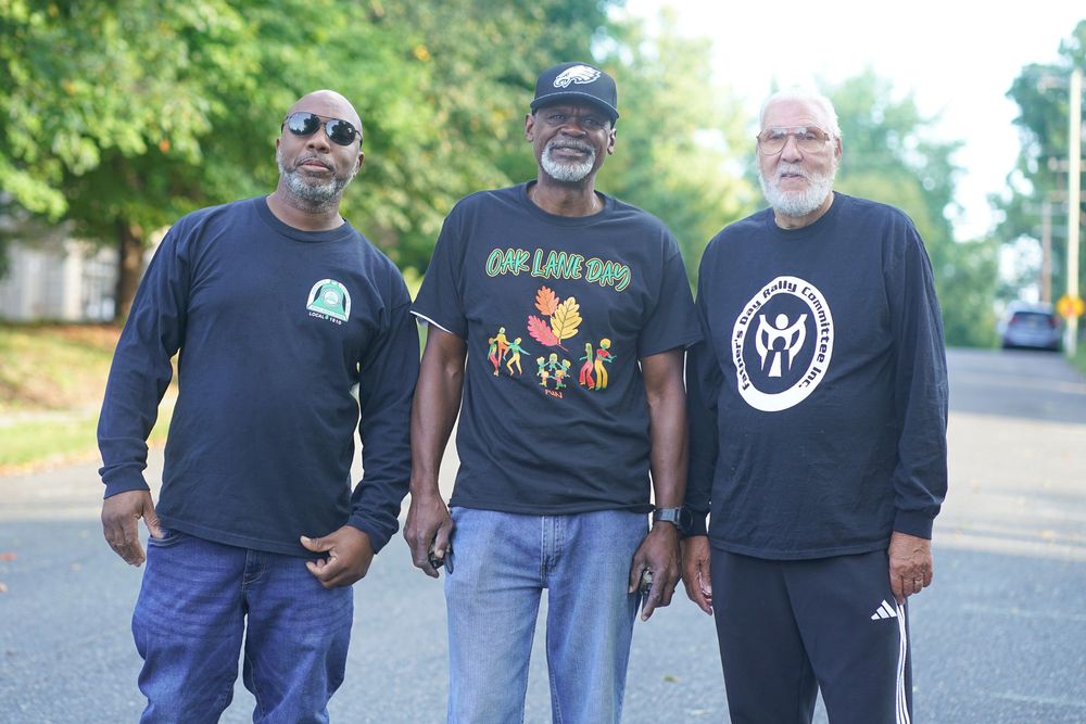 Three men standing together outdoors on a street, wearing casual long-sleeve shirts at a community event.