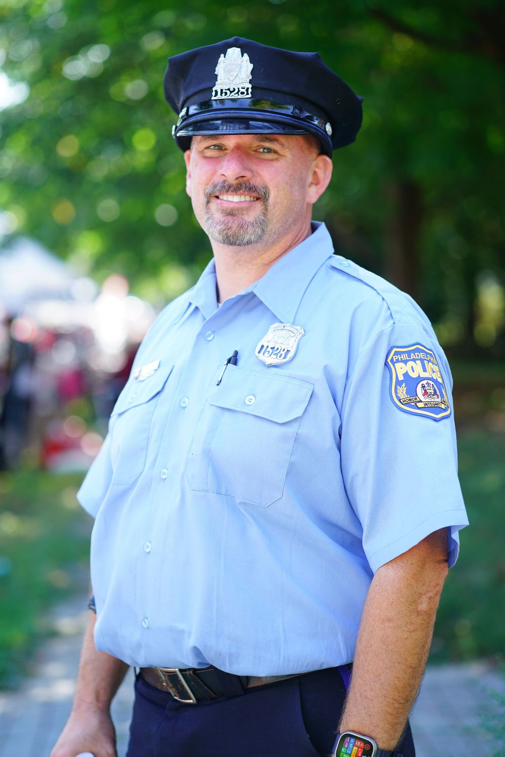 Smiling male Philadelphia police officer in uniform standing outdoors at a community event.