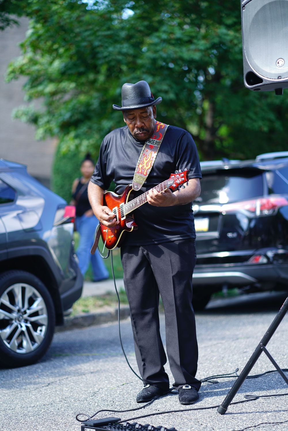 A Black musician in a black hat and outfit plays an electric guitar outdoors at a street performance.