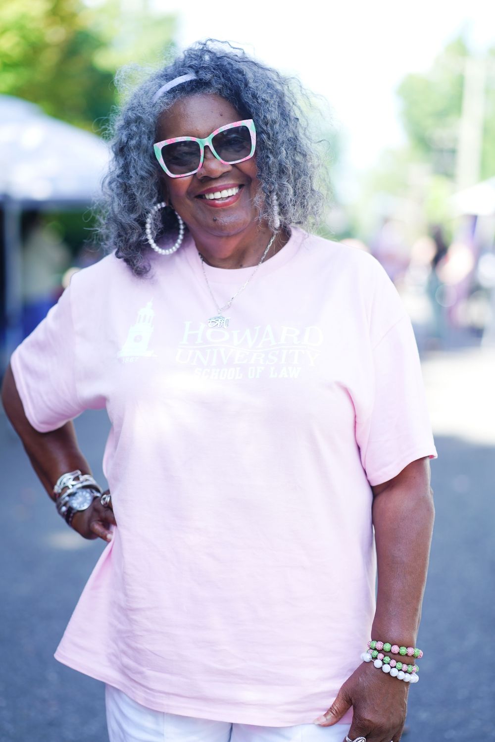 A smiling older Black woman wearing a pink Howard University t-shirt and white sunglasses poses outdoors.