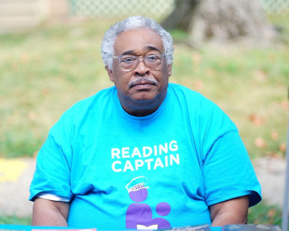 An older man wearing a bright turquoise Reading Captain t-shirt sits outdoors in a park setting.