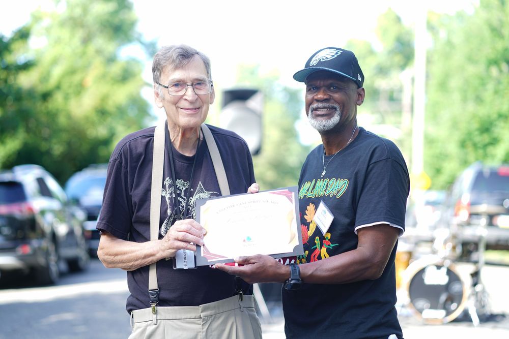 Two men, one elderly white and one Black, stand together outdoors holding a certificate or award.