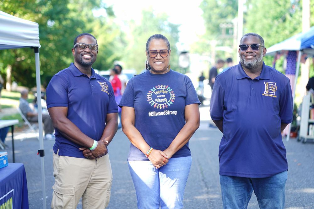 Three smiling people in matching navy blue polo shirts pose together outdoors at a community event.