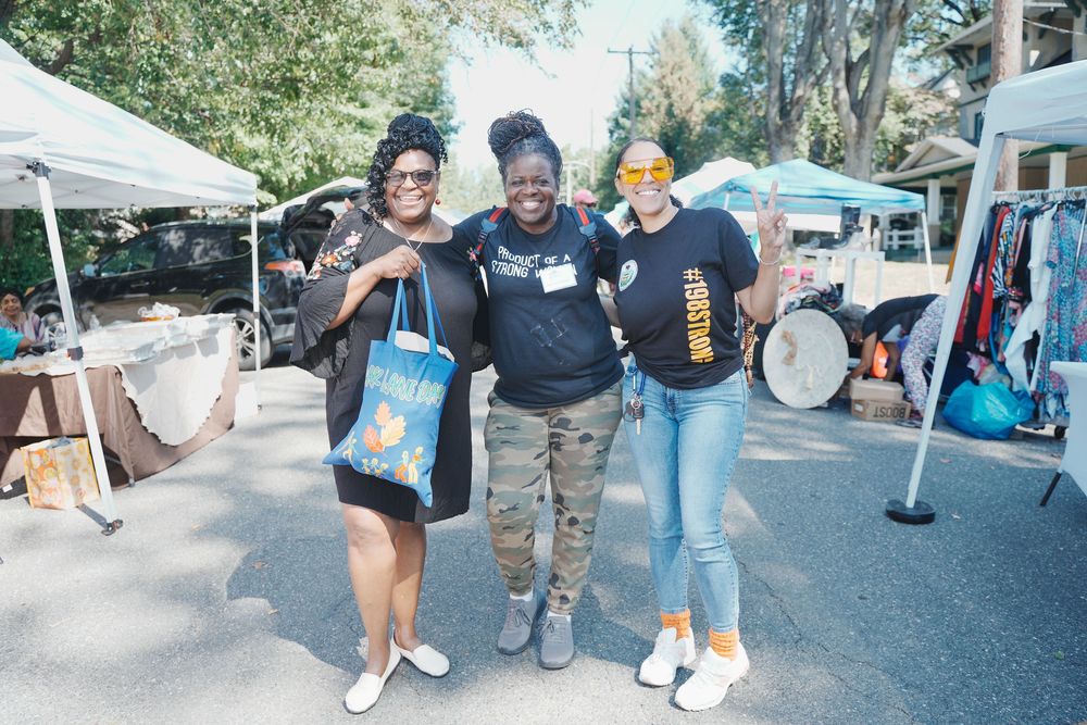 Three women smile and pose together at an outdoor community market event on a sunny day.