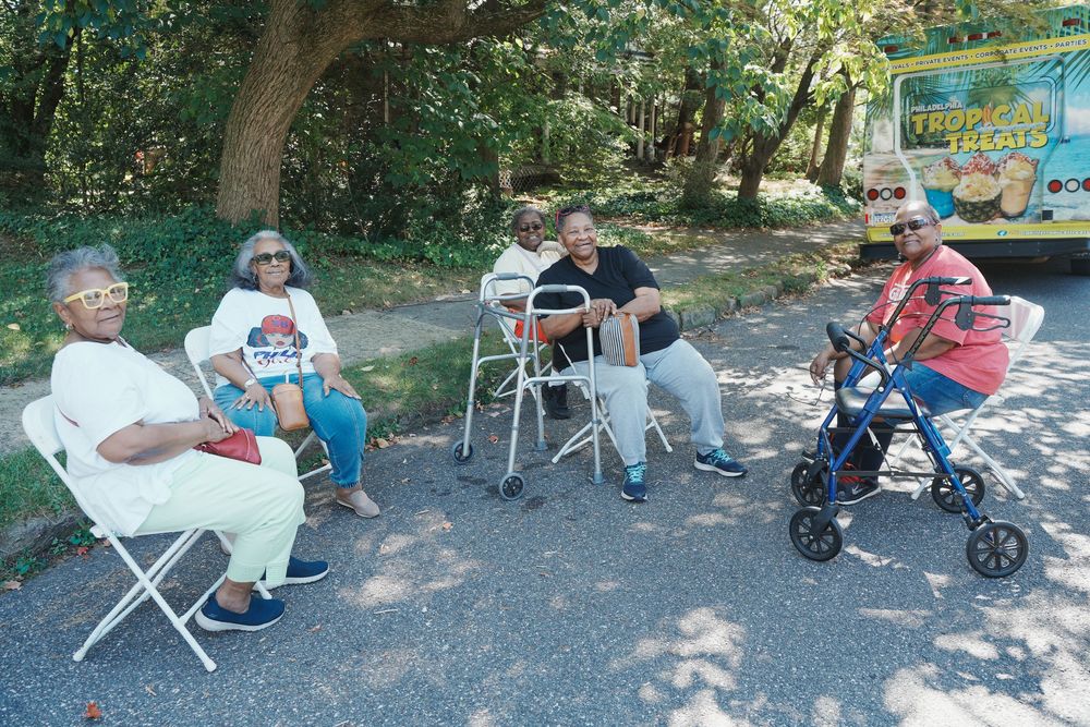 Group of five people relaxing in chairs outdoors near a food truck, some with mobility aids.