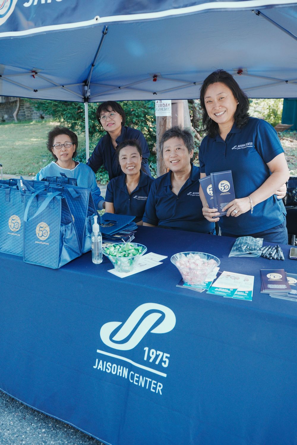 Five women in navy Jaisohn Center uniforms staffing an information booth at an outdoor community event.