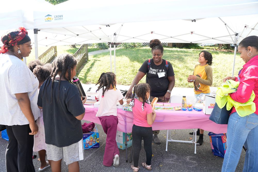 A group of children and adults gather around a pink activity table under a canopy at an outdoor event.