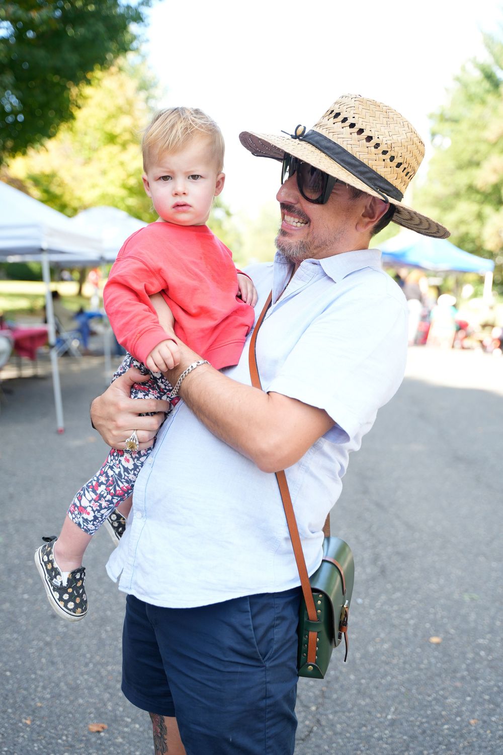 A smiling man in a straw hat and sunglasses holds a toddler in a red shirt at an outdoor market.