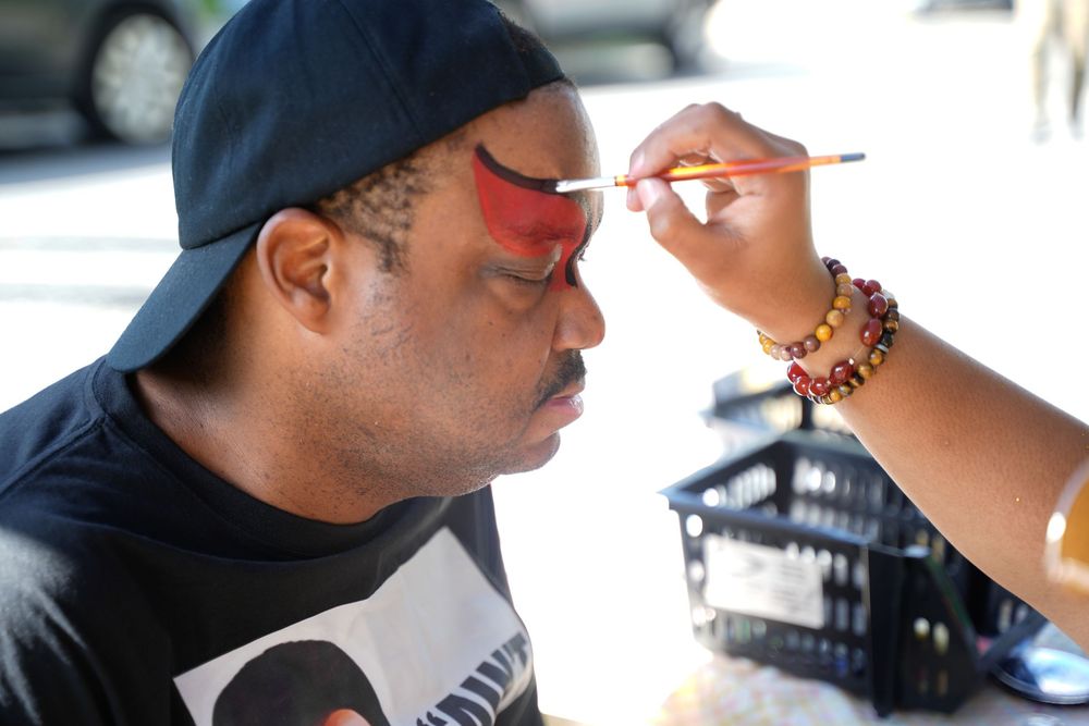 A man in a black cap gets his face painted with red and orange colors by an artist with beaded bracelets.