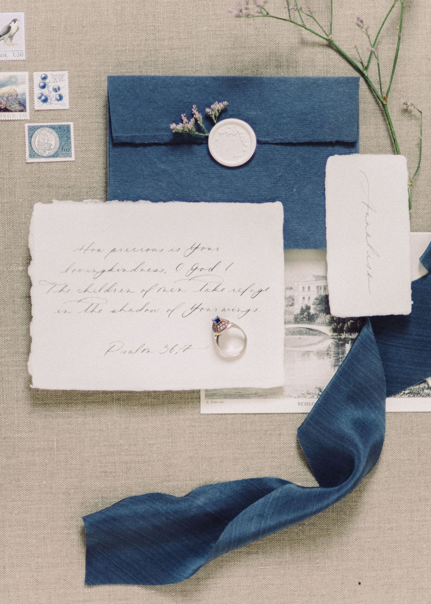 Elegant blue wedding invitation suite with wax seal, calligraphy, silk ribbon, and diamond ring on linen background.