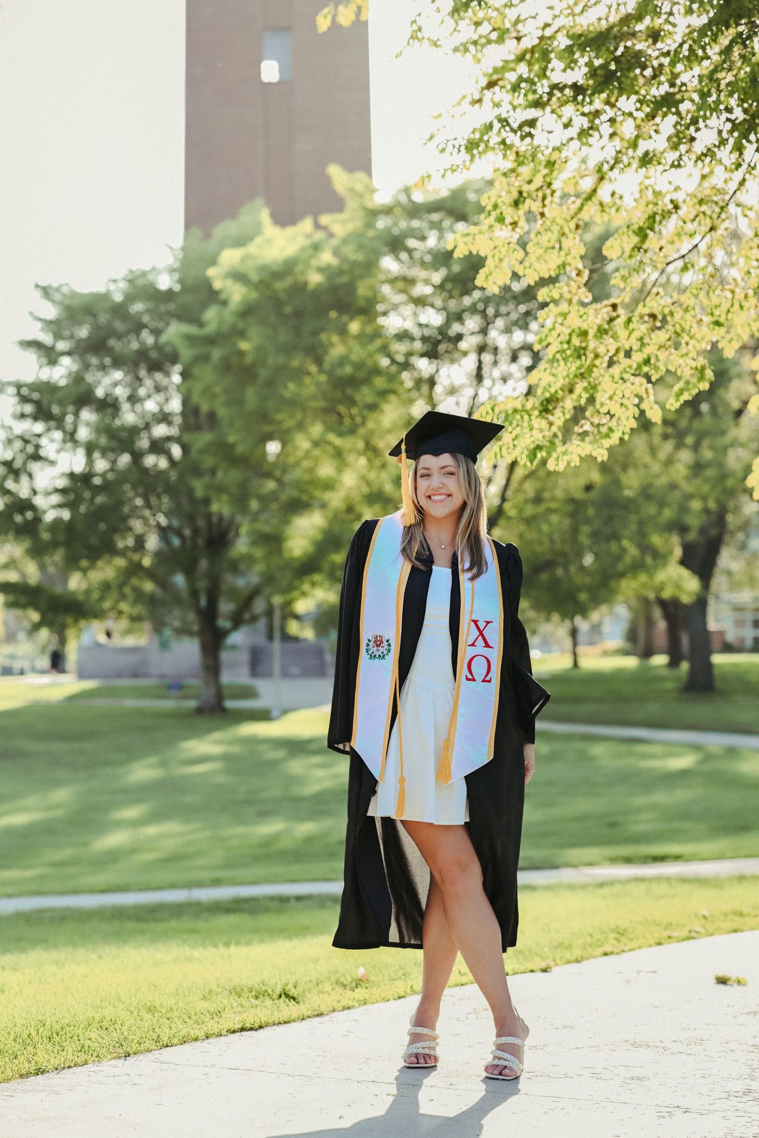 Senior girl in cap and gown photographed outdoors on campus with bright natural light and timeless graduation portrait editing.