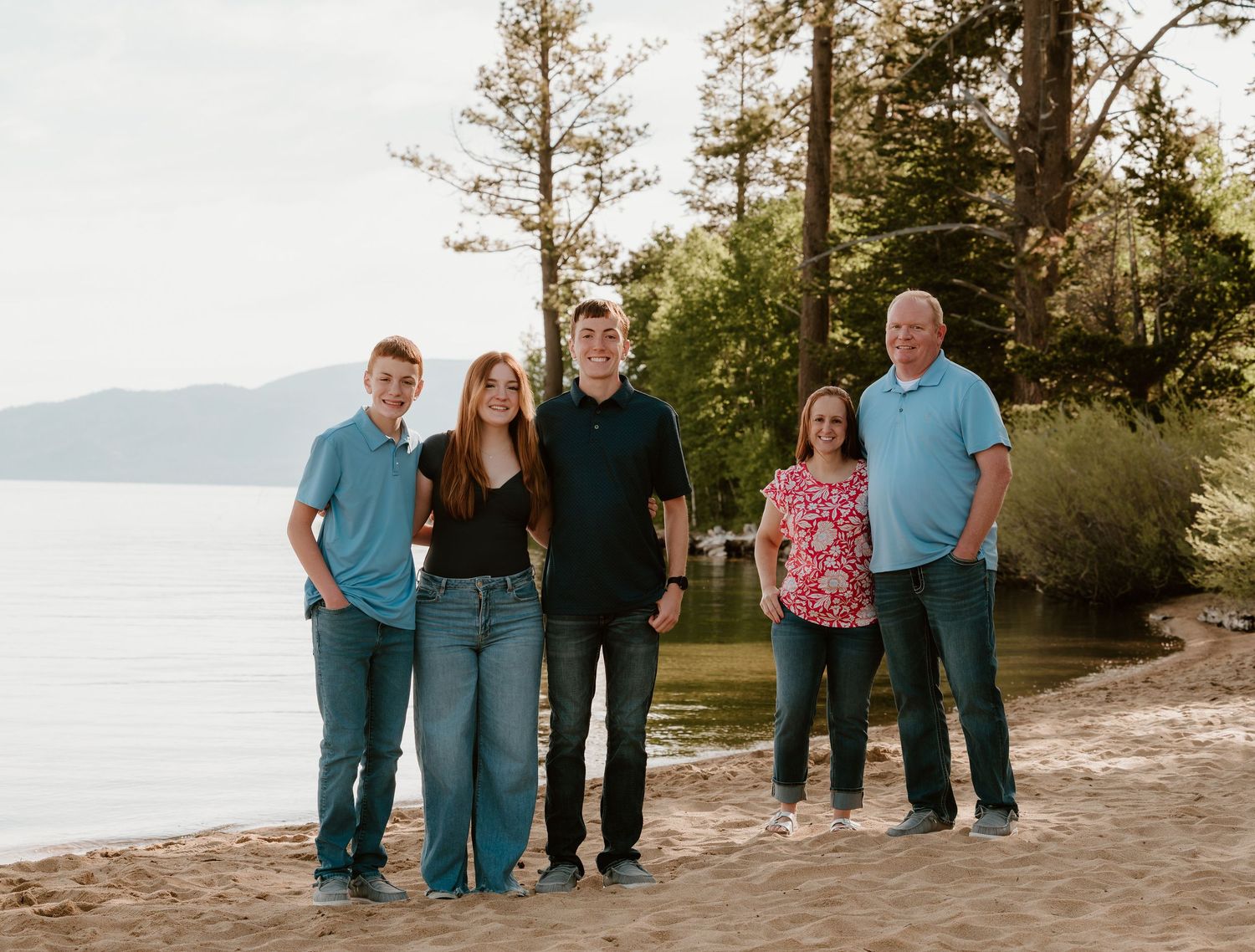 Family of five posing together on a sandy beach near a lake surrounded by tall pine trees during golden hour.