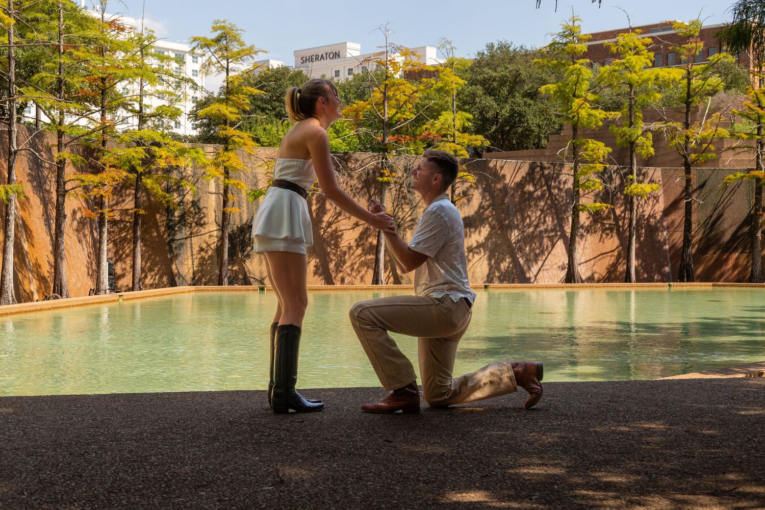 Man proposing to woman on one knee beside a pool, surrounded by trees in an outdoor setting.