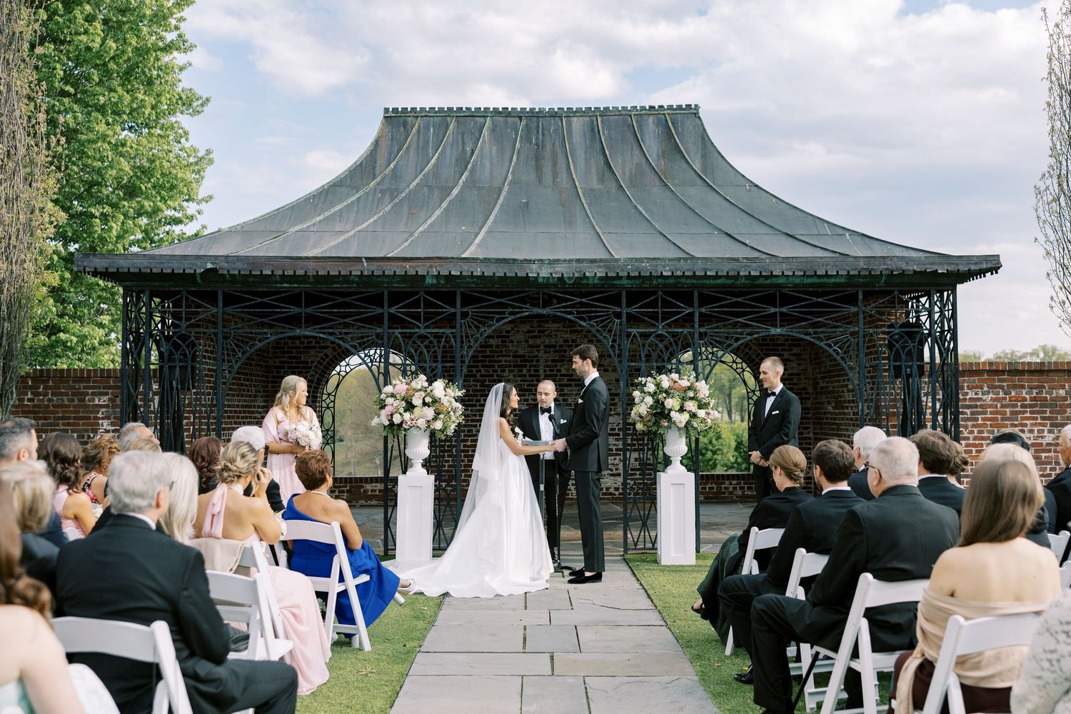 Elegant outdoor wedding ceremony in front of ornate black iron gazebo with floral arrangements and white chairs.