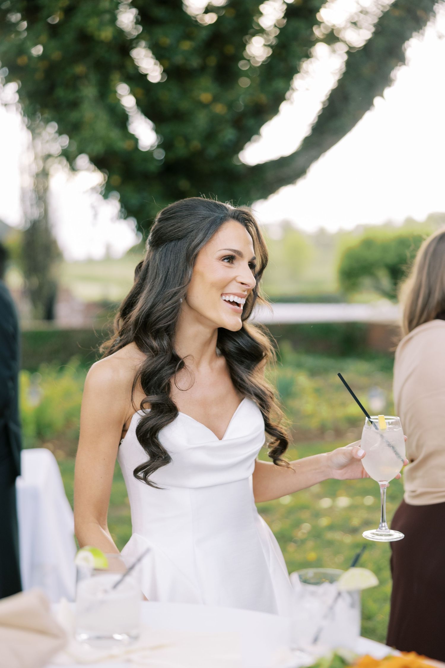 Smiling bride in white strapless gown holding wine glass at outdoor wedding reception.