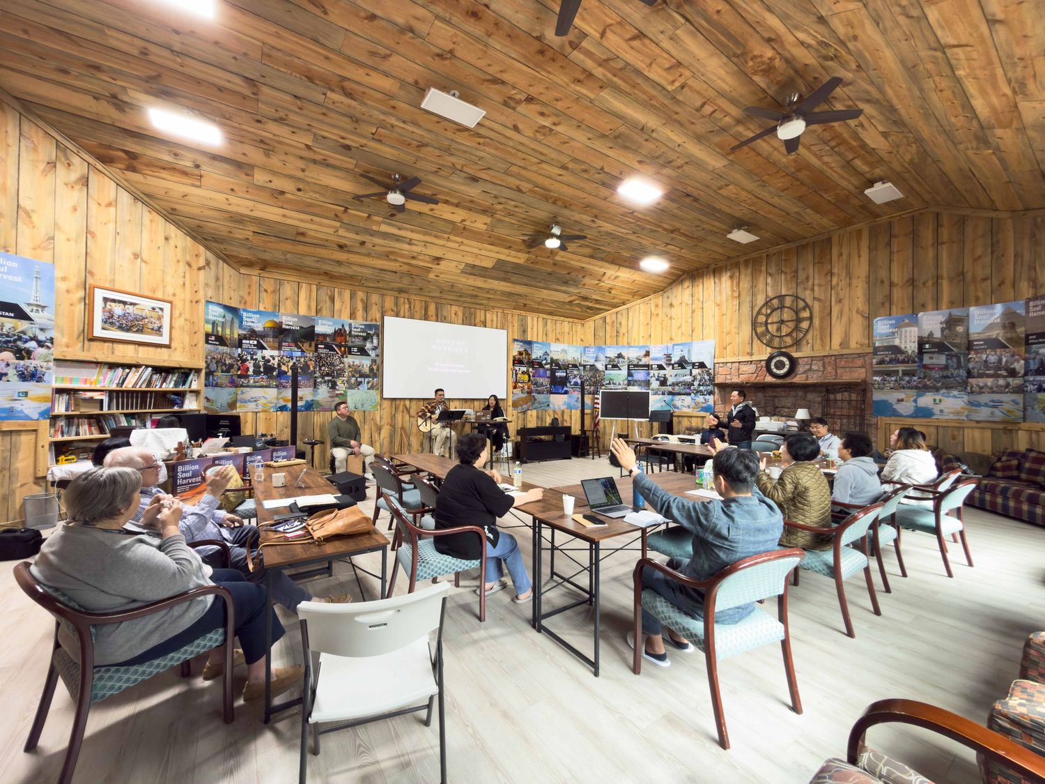 Group of adults attending a workshop in a rustic wood-paneled room with photos on walls and a whiteboard presentation.
