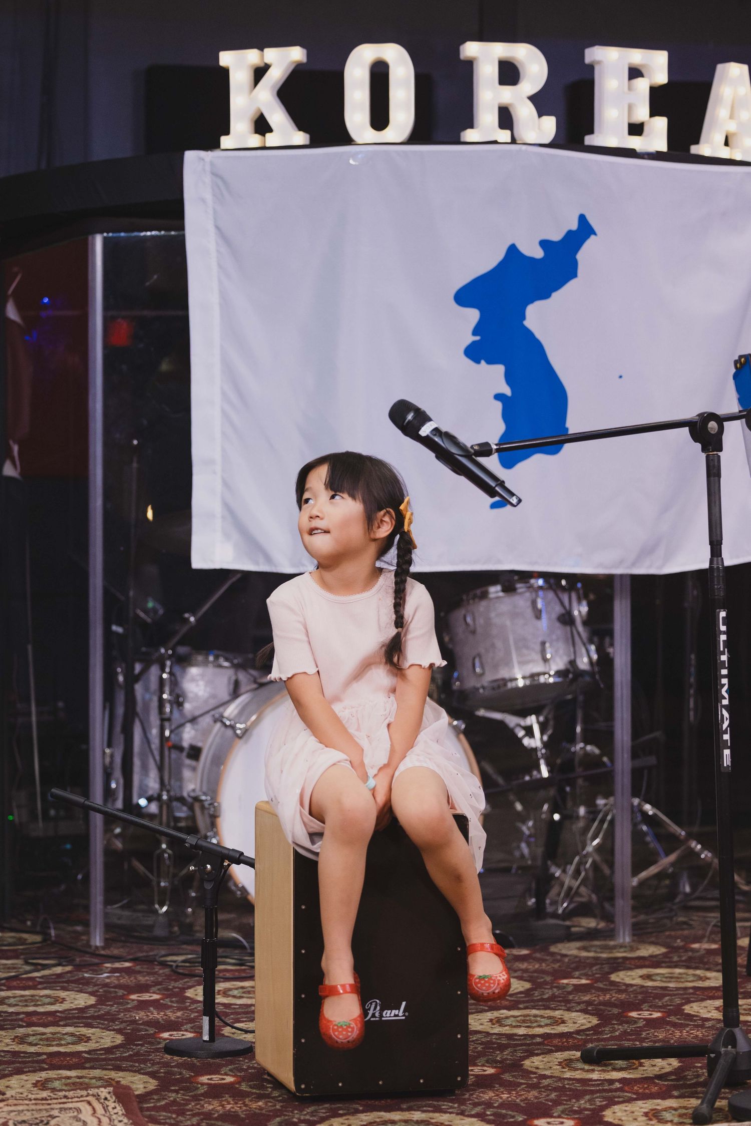 Young girl sitting on a cajon drum box on stage at a Korea cultural event with flag backdrop.