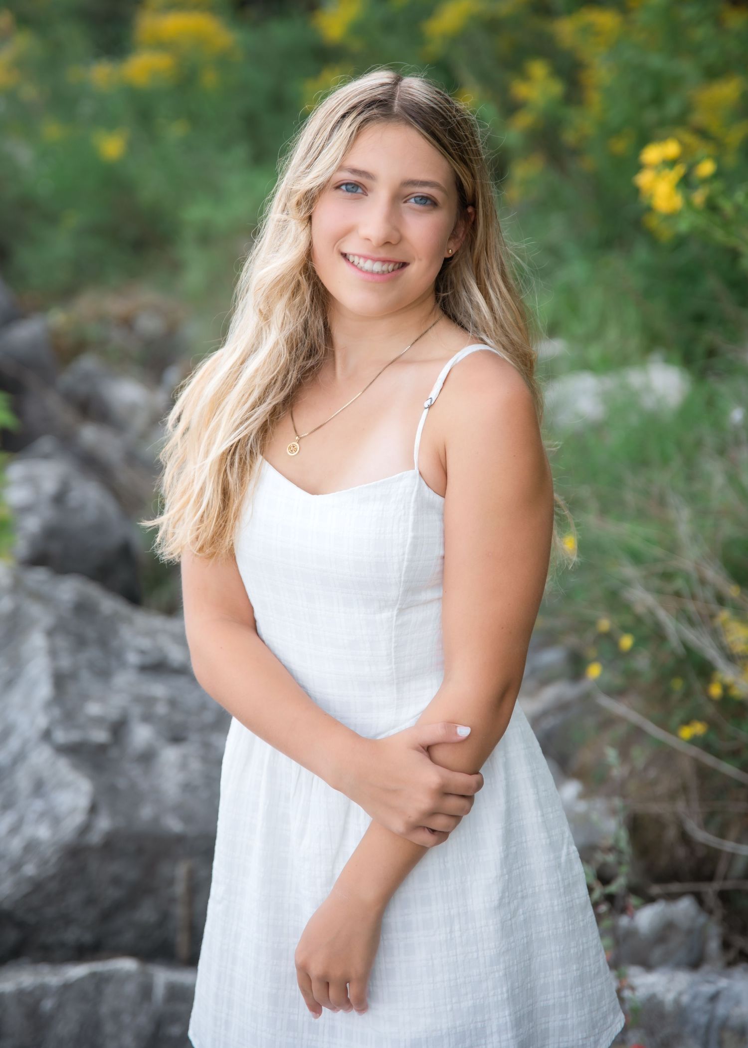 Young woman with blonde hair wearing a white sundress poses outdoors among rocks and yellow wildflowers.