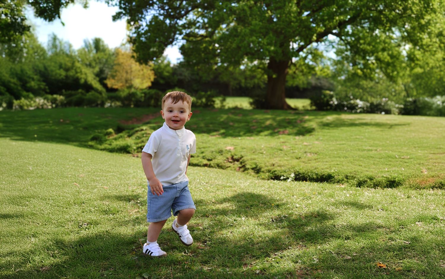 A happy toddler boy in white shirt and denim shorts runs on lush green grass in a sunny park.