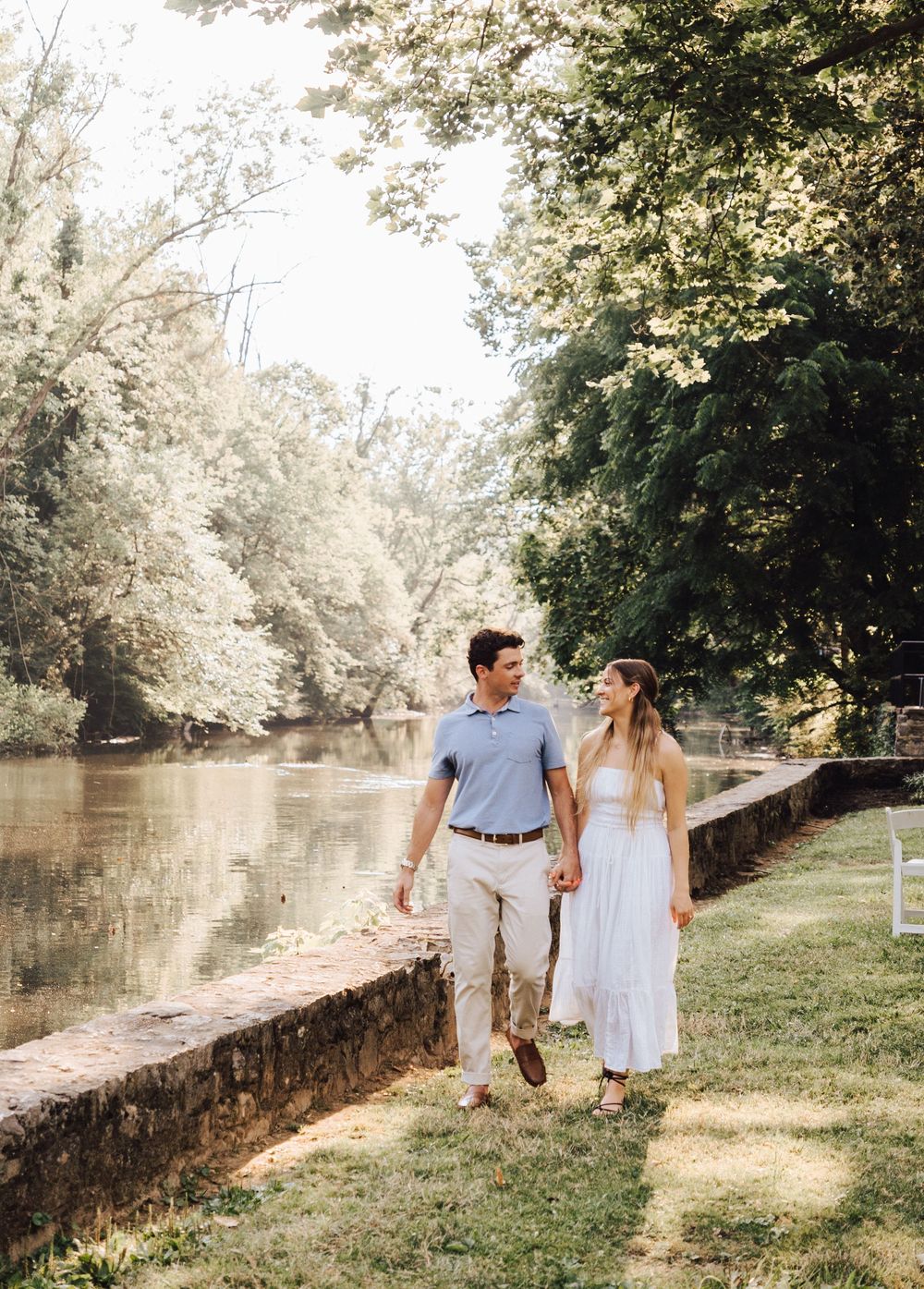 Couple holding hands walking along a scenic riverside path surrounded by lush green trees on a sunny day.
