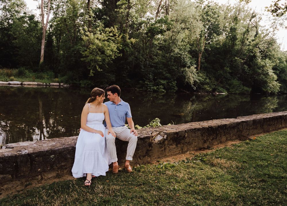 Couple in wedding attire sitting on a stone wall by a serene river surrounded by lush green trees.