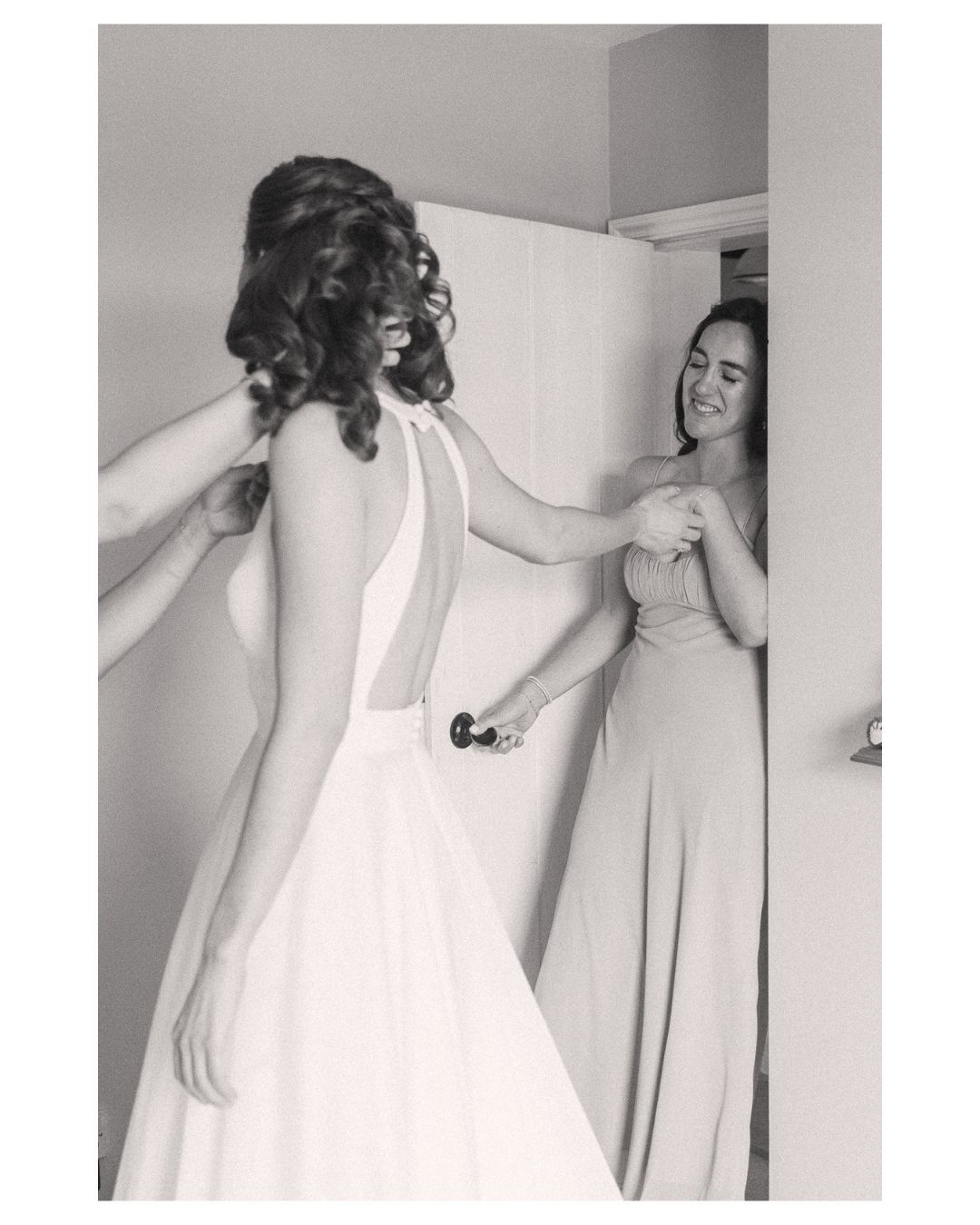 Black and white photo of a bride in a backless gown being helped by a smiling bridesmaid.