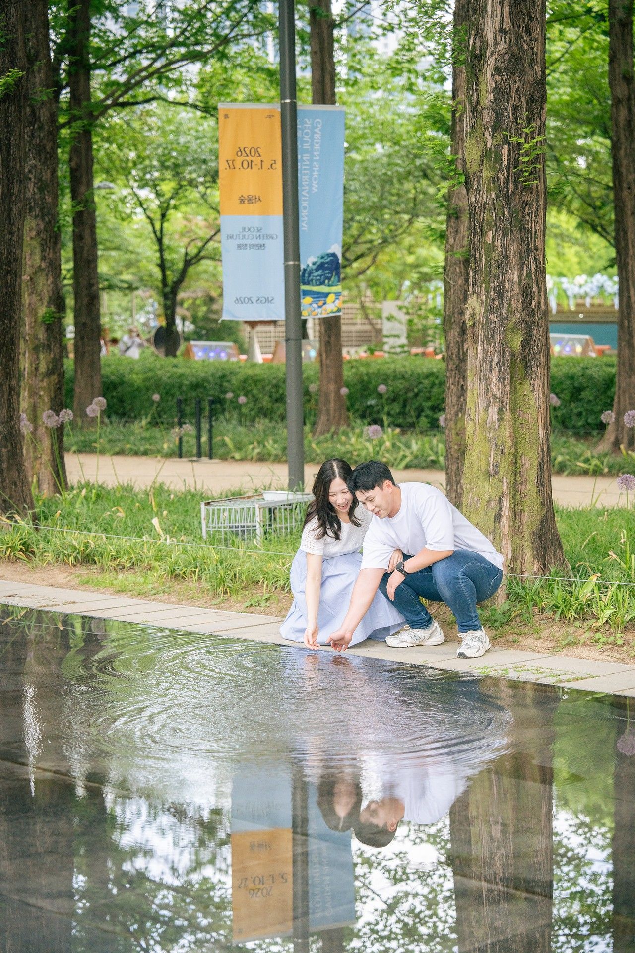 Couple crouching by a reflective pool in a lush green park, surrounded by tall trees and colorful banners.
