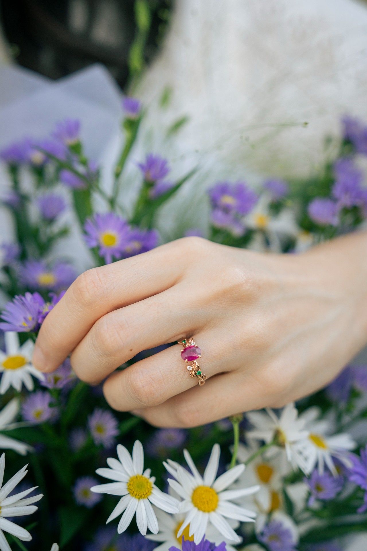 Hand wearing gold ring with pink gemstone resting on bouquet of purple wildflowers and white daisies.