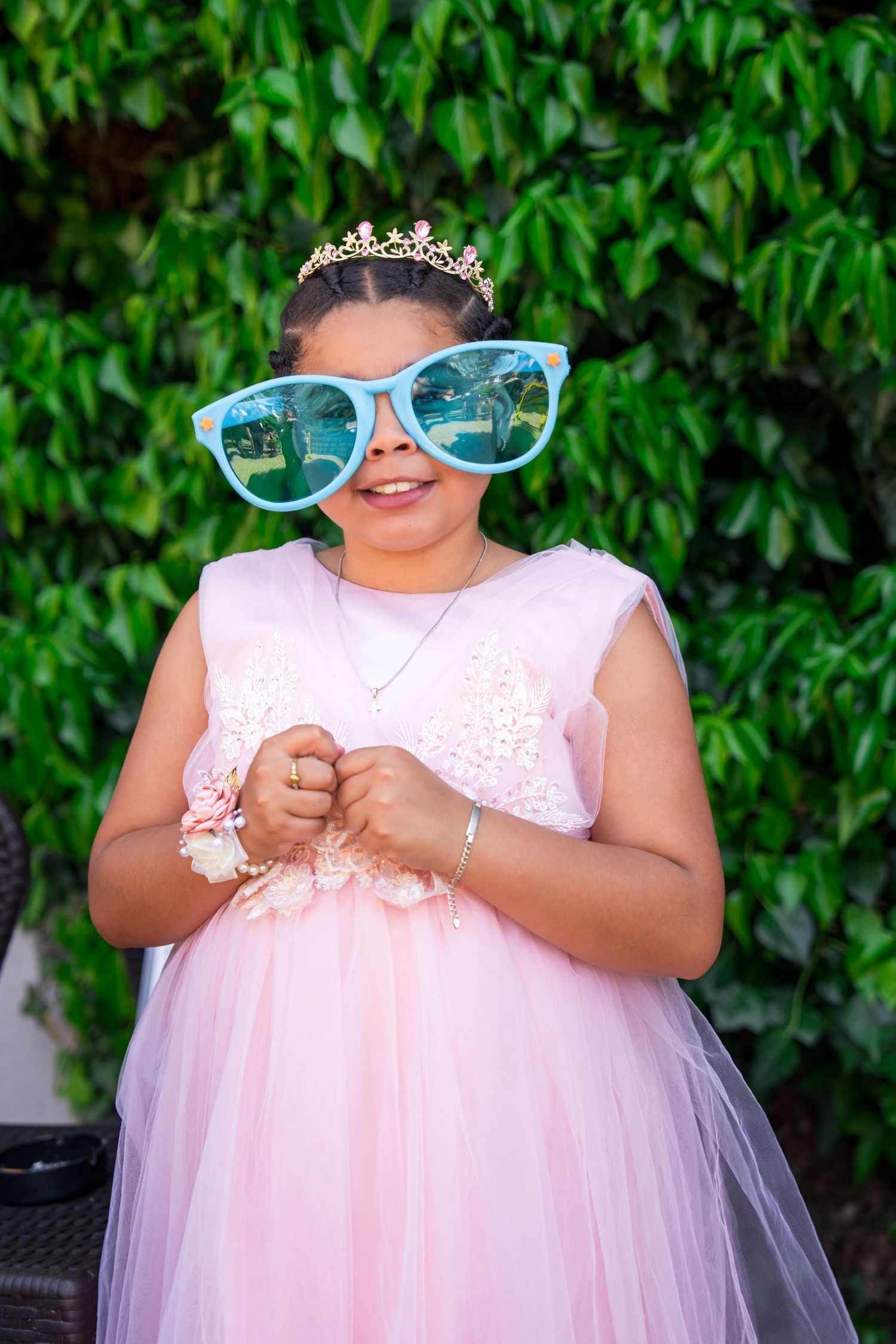 Young girl in pink princess dress and tiara wearing oversized blue sunglasses, smiling outdoors.