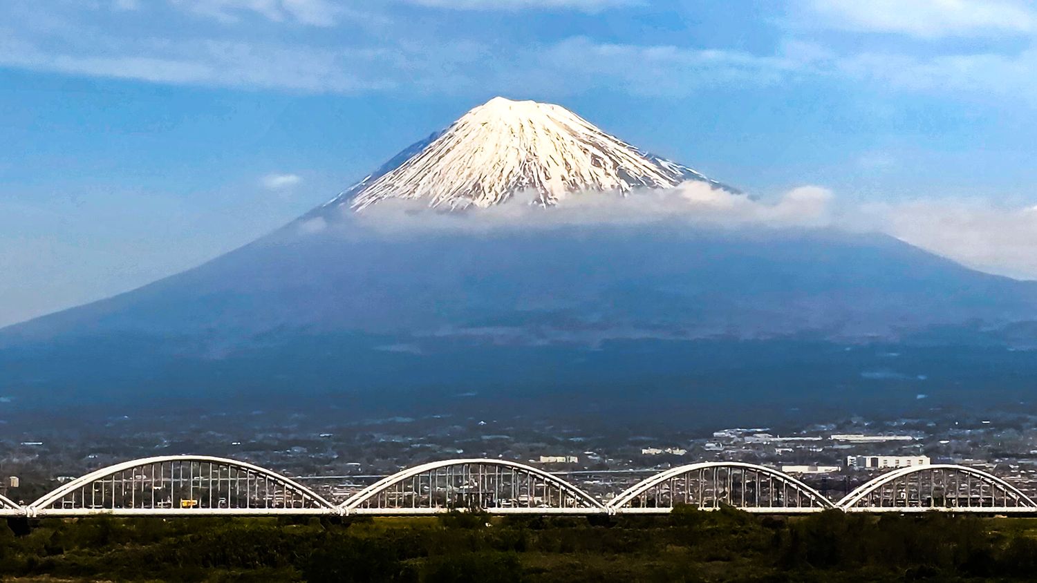 Snow-capped Mount Fuji towers over a bridge and cityscape under a partly cloudy blue sky in Japan.