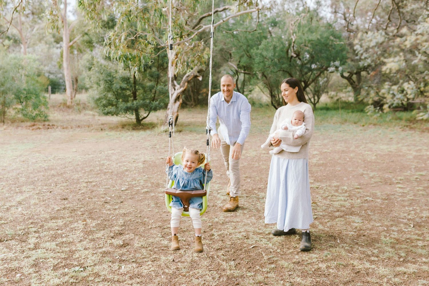 Family enjoying outdoor park with toddler on rope swing, parents smiling, mother holding newborn baby, surrounded by trees.