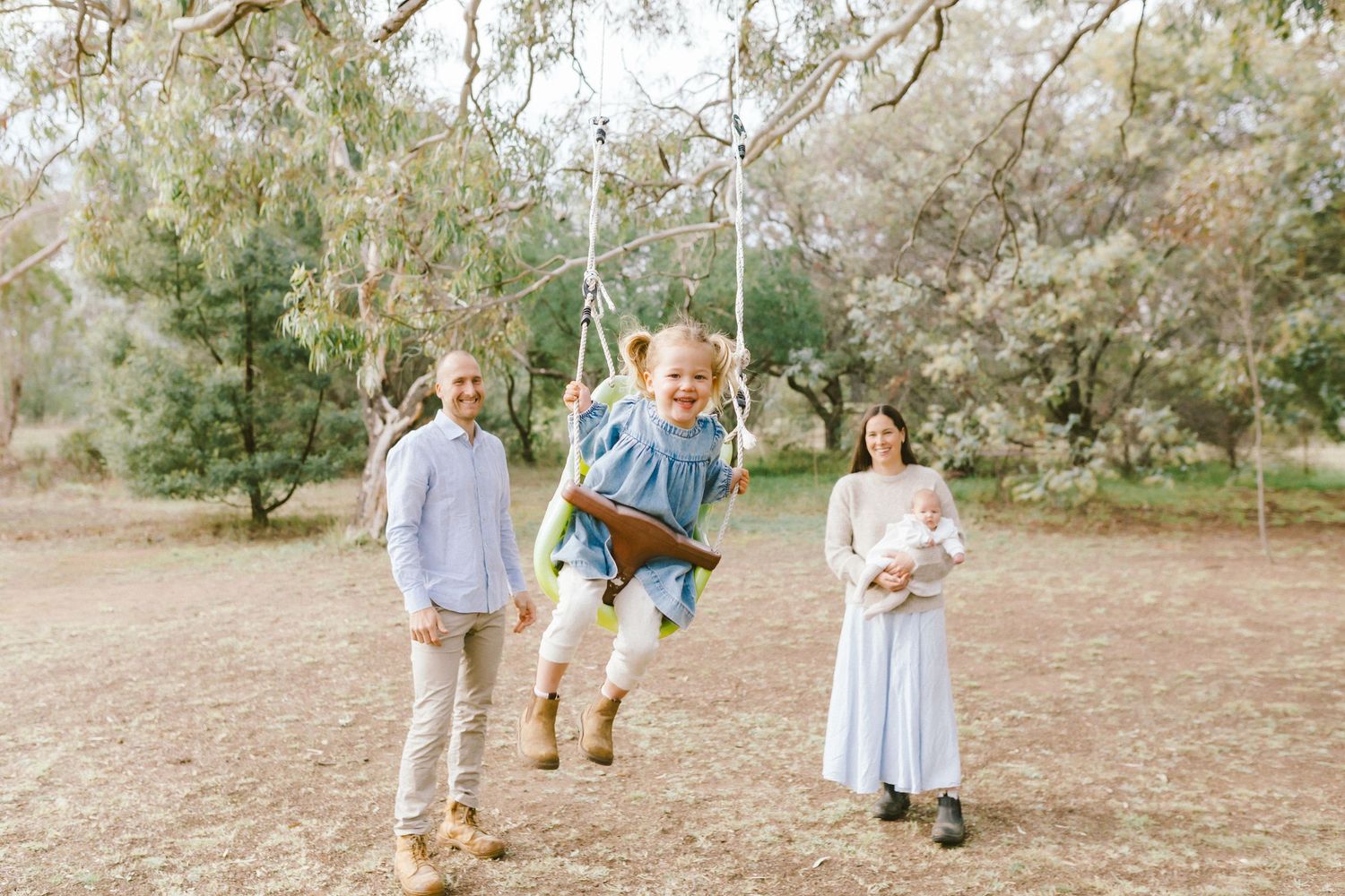 Family photo with child joyfully swinging outdoors while parents stand nearby holding a newborn baby in a park.