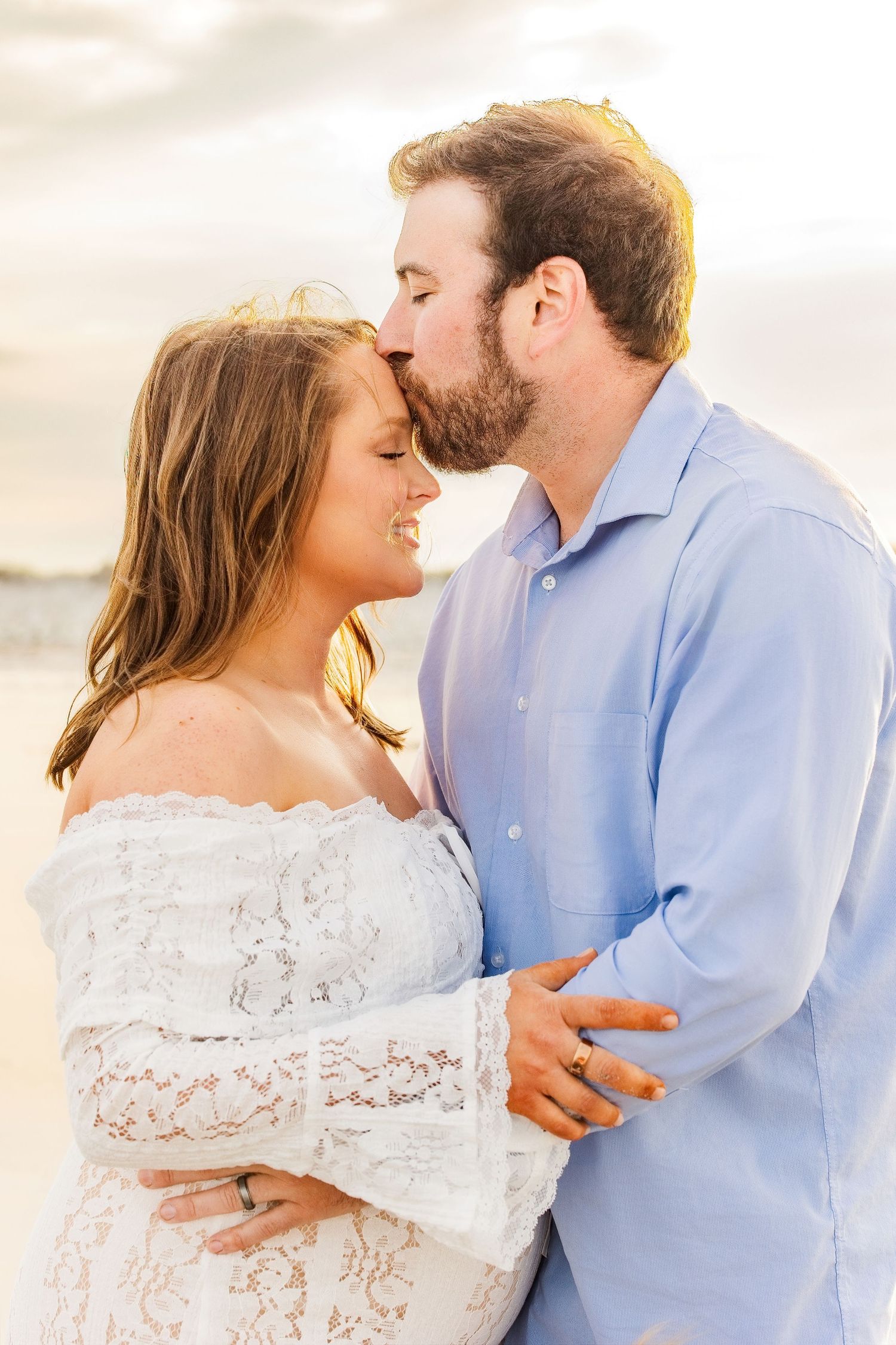 A couple shares a tender moment on the beach at sunset, man kissing woman's forehead as she smiles warmly.