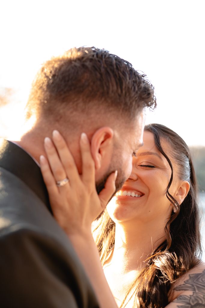 A joyful bride with a ring on her finger tenderly holds her groom's face as they share a romantic moment in golden light.