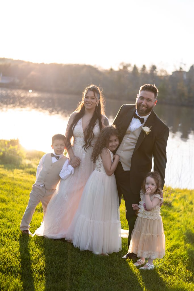 Bride and groom pose with three young children on a sunlit lawn by a lake during an outdoor wedding ceremony.