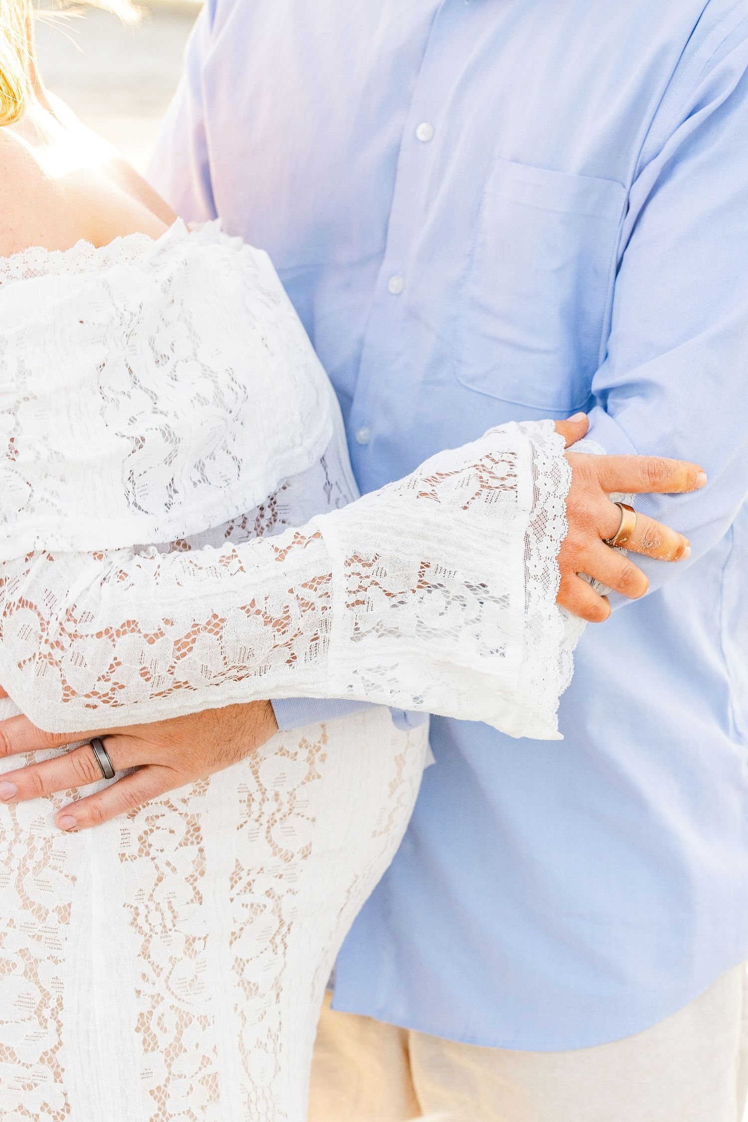 Couple embracing, woman in white lace dress and man in light blue shirt, both wearing wedding rings.