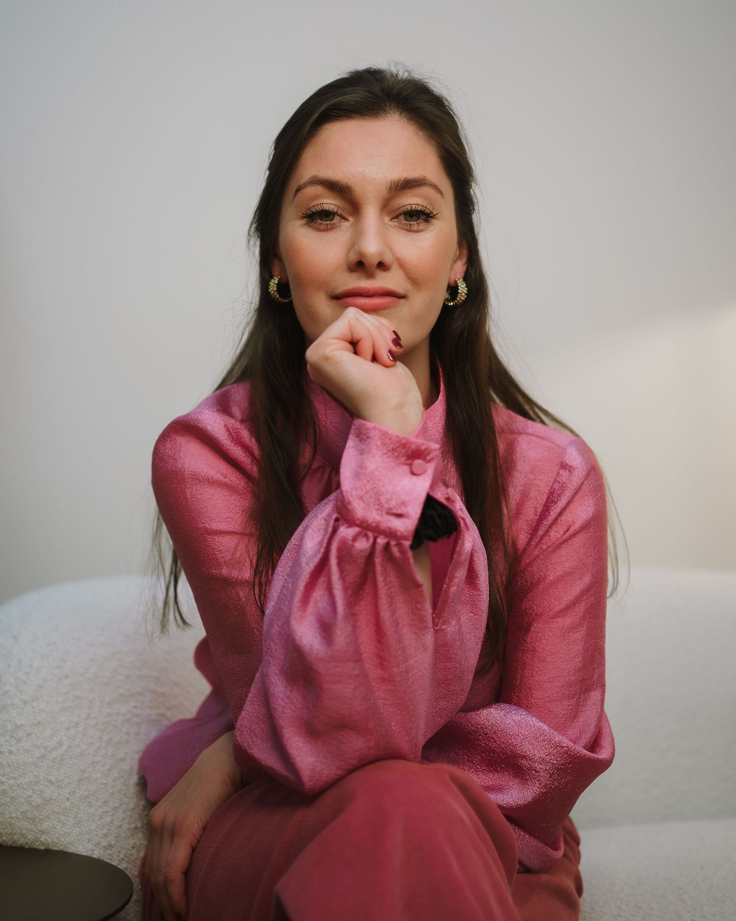 Woman in pink silk blouse seated gracefully, posing with hand on chin against a white background.